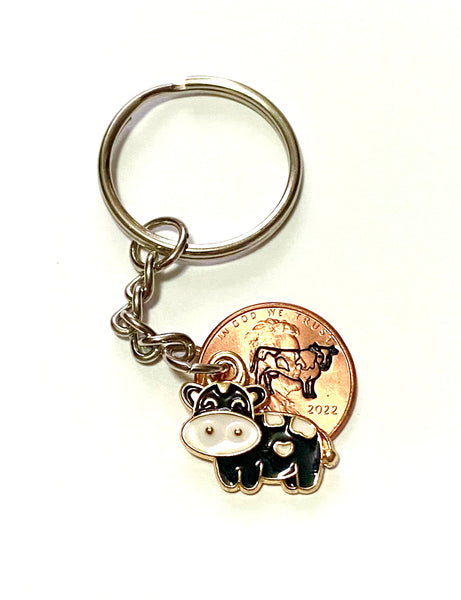 A black cow charm with a big pink nose that has a white heart stamp on it's belly with a hand stamped cow engraved on a penny.