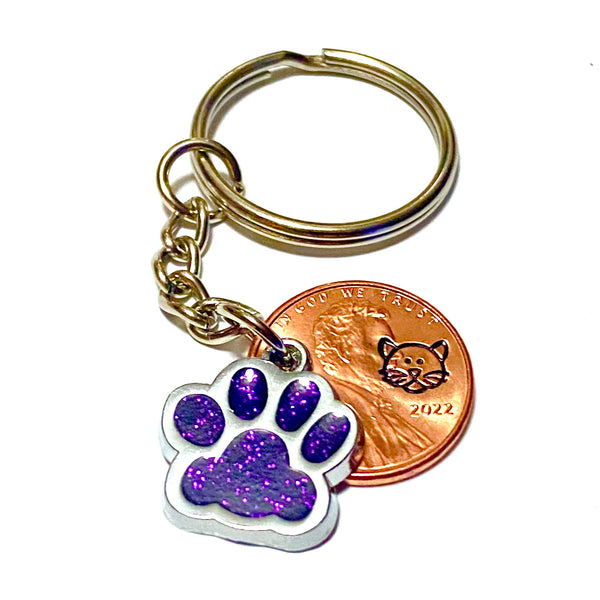 A purple glitter charm of a cat's paw with a hand stamped penny with a cat's face on a lucky penny keychain.