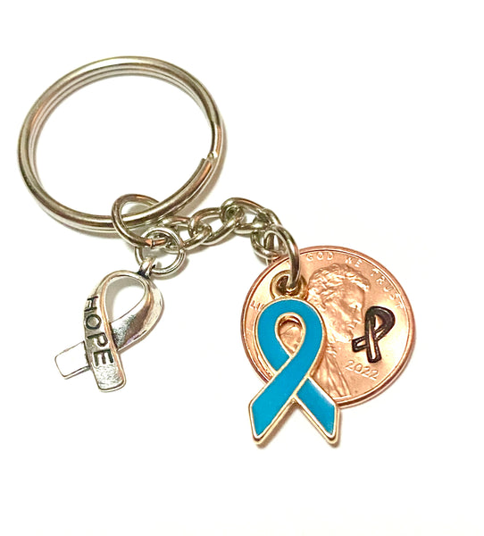 A blue awareness ribbon along with a silver HOPE charm attached to a Lincoln Cent that is hand stamped with a detailed ribbon design to make a great Lucky Penny Keychain.