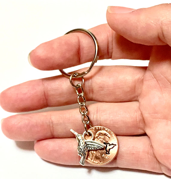 A Silver Hummingbird Charm attached to an engraved Lincoln Cent with a matching design above the date of a Lincoln Cent.
