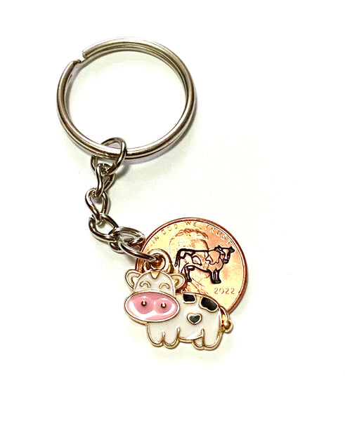 A white cow charm with a big pink nose that has a black heart stamp on it's belly with a hand stamped cow engraved on a penny.