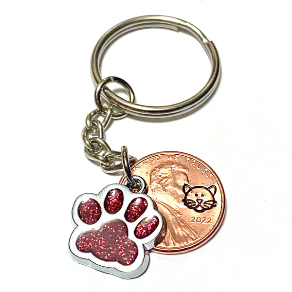 A cat face cat paw print glitter charm on a 3" keychain. We engrave your lucky penny with a cat face stamp and match it with a glitter charm.