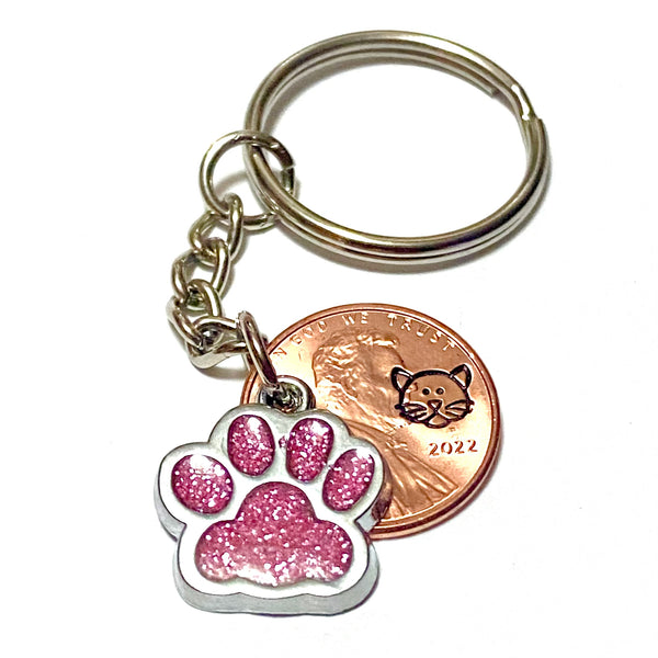 A pink glitter charm of a cat's paw with a hand stamped penny with a cat's face on a lucky penny keychain.
