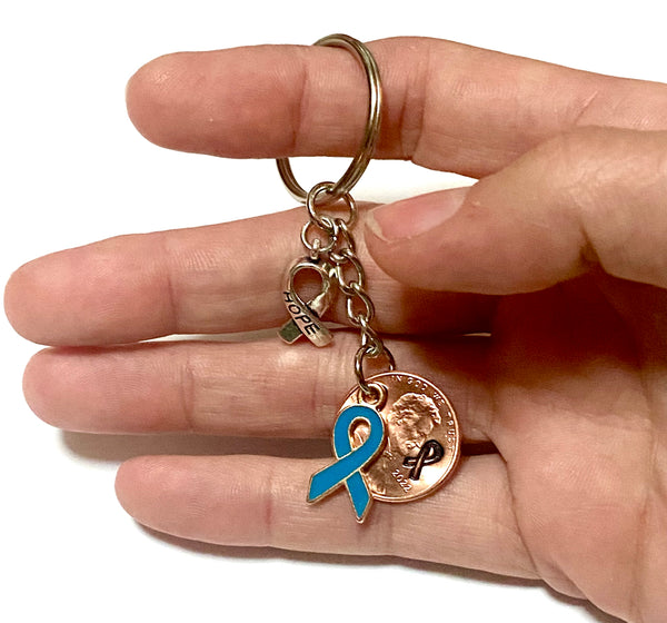 A blue awareness ribbon along with a silver HOPE charm attached to a Lincoln Cent that is hand stamped with a detailed ribbon design to make a great Lucky Penny Keychain.