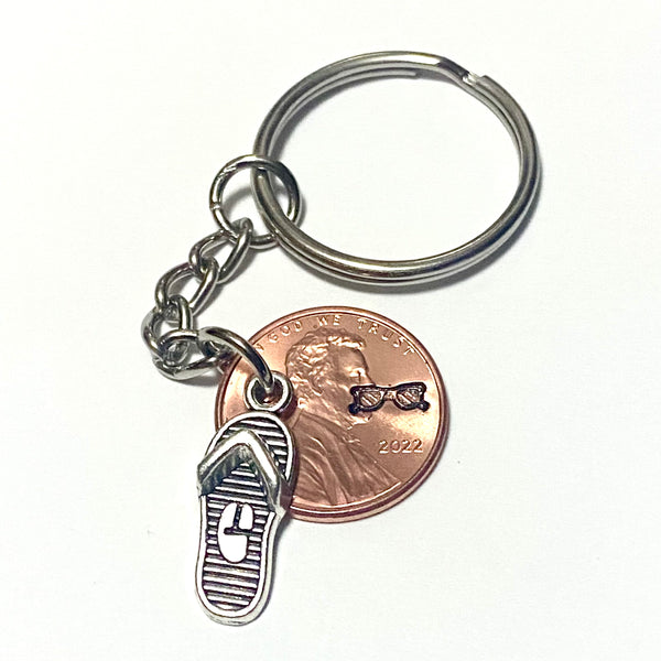 A silver Flip Flop charm attached to a Lincoln Cent with a hand stamped design of a pair of sunglasses engraved above the date.